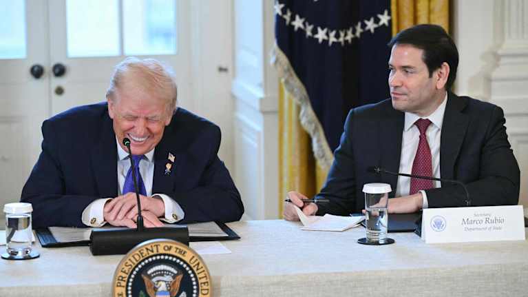Donald Trump sitter vid ett bord och skrattar medan Marco Rubio ser på honom.