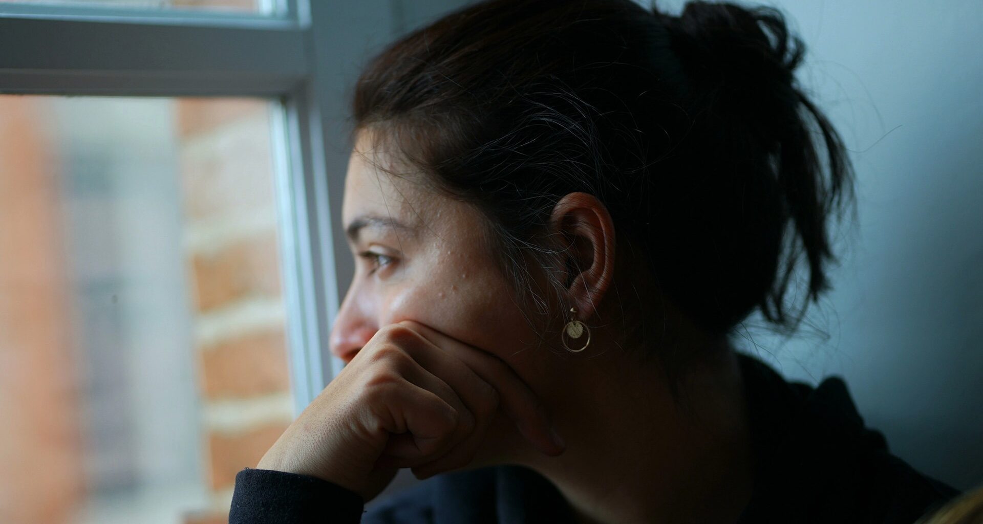 Tankfull kvinna med mörkt hår i en tofs tittar ut genom ett fönster.