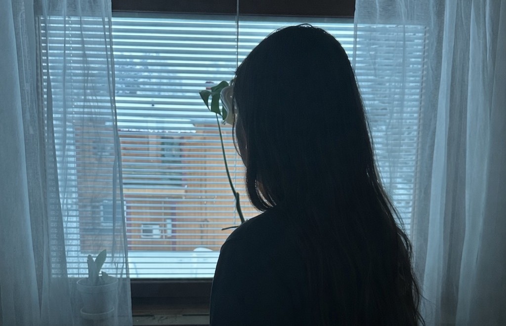 En person med långt hår står inomhus, vänd mot ett fönster med skira gardiner och persienner, och tittar ut på en snötäckt byggnad - en lugn stund då han reflekterar över migration. På fönsterbrädan står en krukväxt.