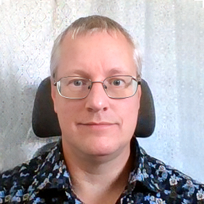 En person med kort ljust hår och glasögon sitter framför en mönstrad ljus bakgrund, klädd i en mörk skjorta med blommönster och tittar direkt in i kameran med ett neutralt ansiktsuttryck.