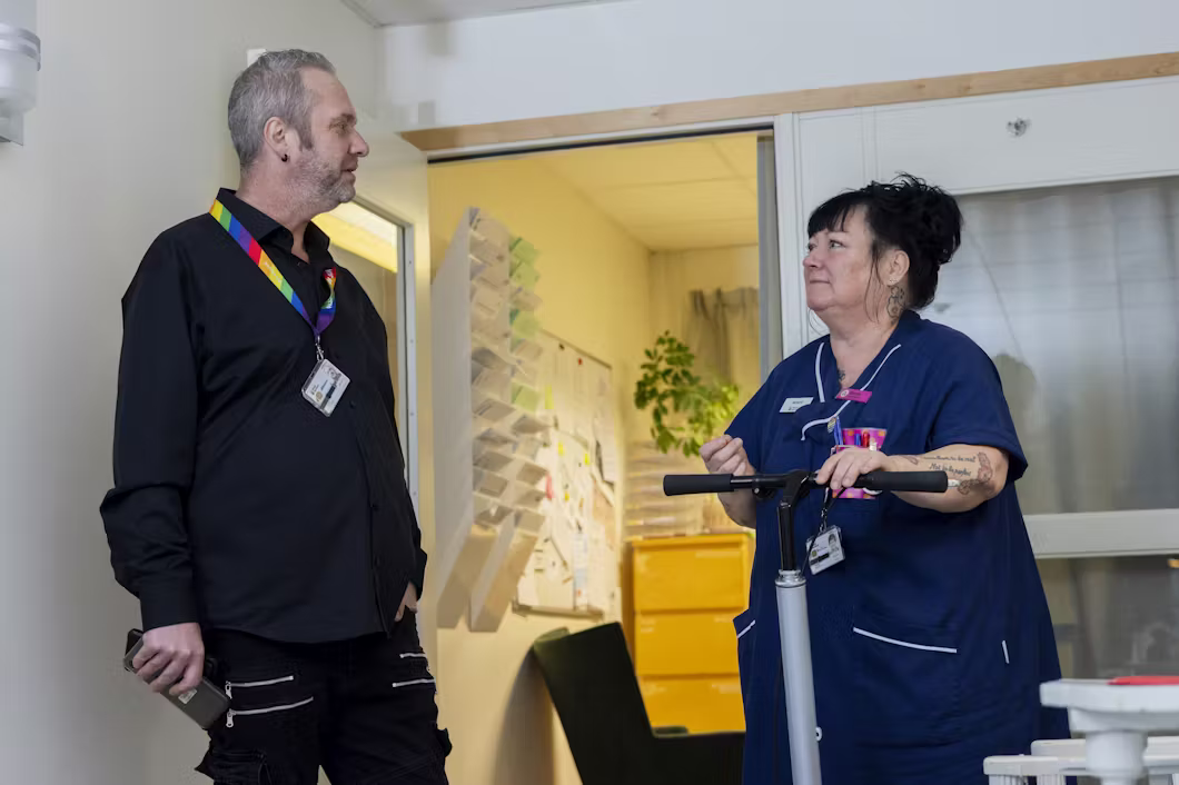 En man i svarta kläder pratar med en kvinna i blå vårdkläder som håller i ett gånghjälpmedel i en vårdmiljö.