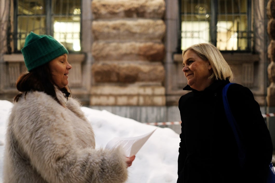 Två kvinnor står utomhus i vinterkläder, den ena håller papper. De samtalar framför en byggnad med snö på marken.
