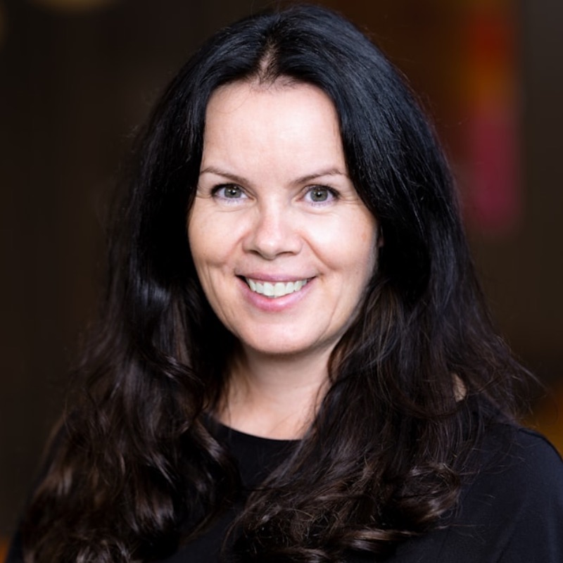 En kvinna med långt, vågigt mörkt hår och ljus hy ler mot kameran. Hon bär en svart topp, medan en suddig bakgrund med mörka och varma toner framkallar en atmosfär där ai tar jobben in i framtiden.