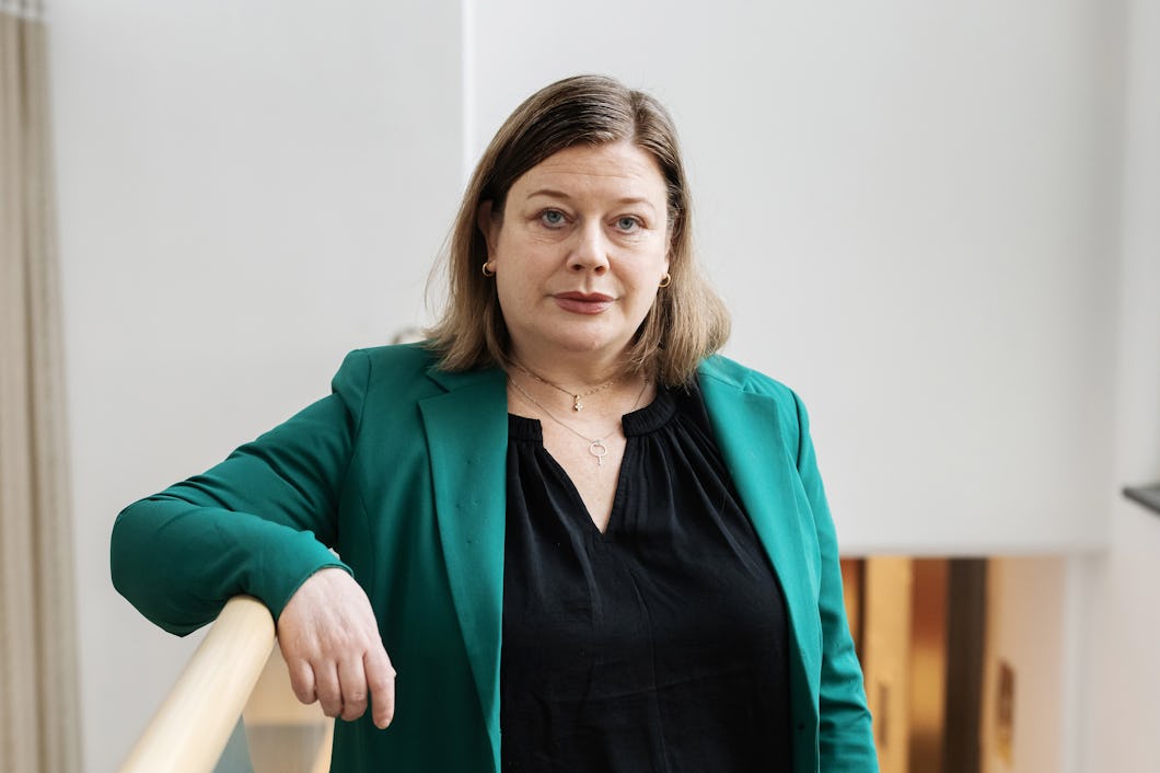 Malin Ragnegård med brunt axellångt hår står inomhus i grön kavaj och svart topp, med handen på ett räcke och ser mot kameran.