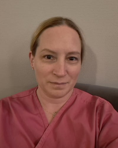 En person med rosa vårdkläder sitter framför en beige vägg och tittar mot kameran med neutral min.