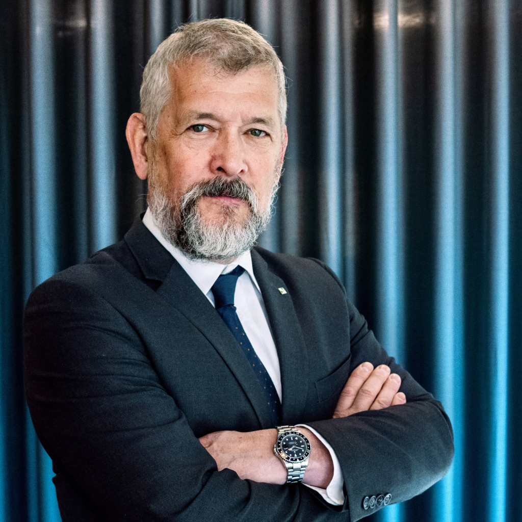 En man med grått hår och skägg klädd i mörk kostym, vit skjorta och blå slips står med armarna i kors framför en bakgrund av mörka vertikala gardiner.