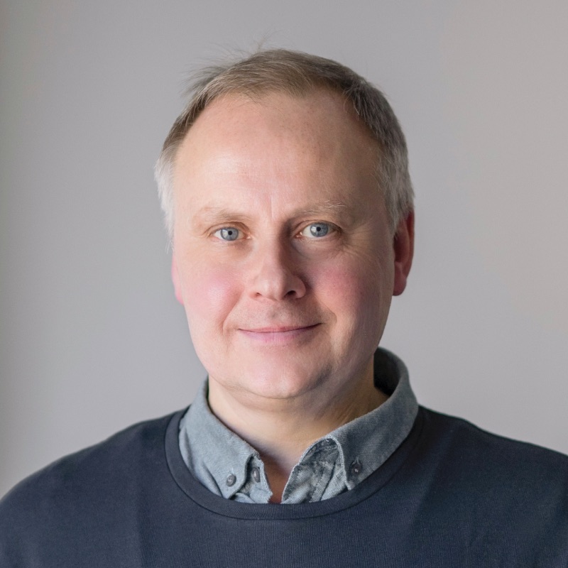 En medelålders man med kort, ljust hår och blå ögon vänder sig mot kameran. Han bär en grå skjorta med krage under en mörk tröja. Den enfärgade, ljusa bakgrunden antyder subtilt en framtid där AI tar jobben endrer arbeidslivet.