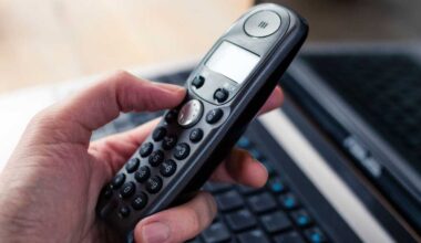 Hand holding a black cordless telephone with a laptop at the background