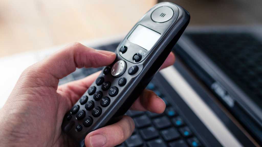 Hand holding a black cordless telephone with a laptop at the background