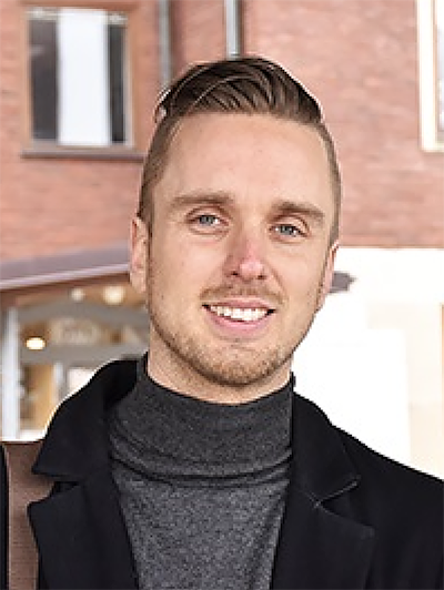 Simon Elfgren med kort, ljusbrunt hår står utomhus framför en tegelbyggnad. Han bär en mörk polotröja och rock och ler mot kameran.