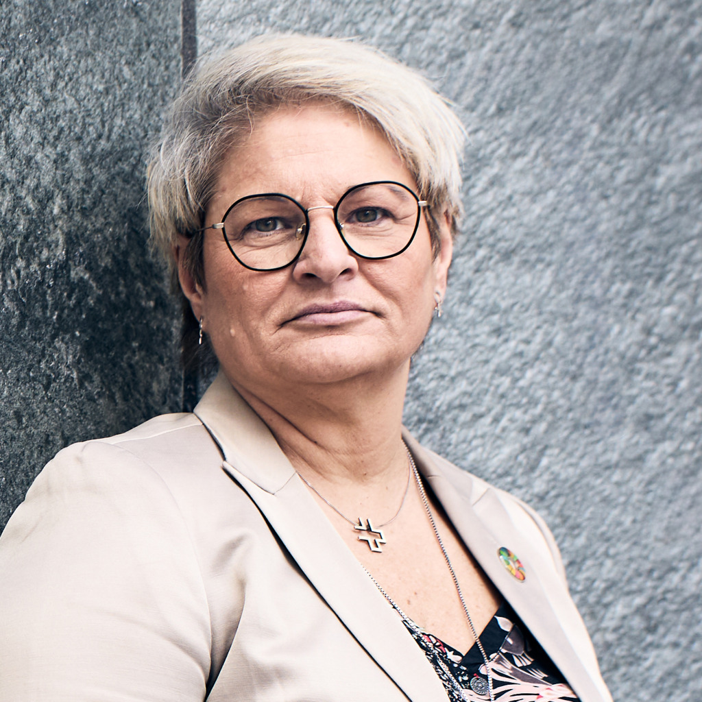 En person med kort blont hår och runda glasögon, klädd i beige kavaj och mönstrad topp, står mot en grå vägg med textur och tittar rakt in i kameran med ett neutralt ansiktsuttryck.