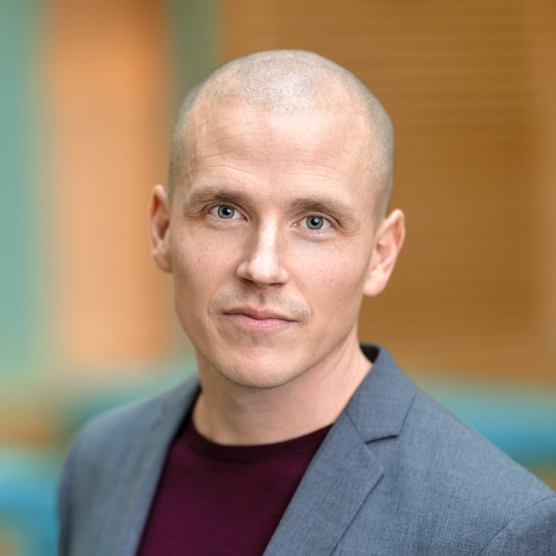 En person med rakat huvud, grå kavaj och mörkbrun skjorta tittar in i kameran med ett neutralt uttryck, som om han funderar på om ai tar jobben. Bakgrunden är mjukt suddig med varma toner.