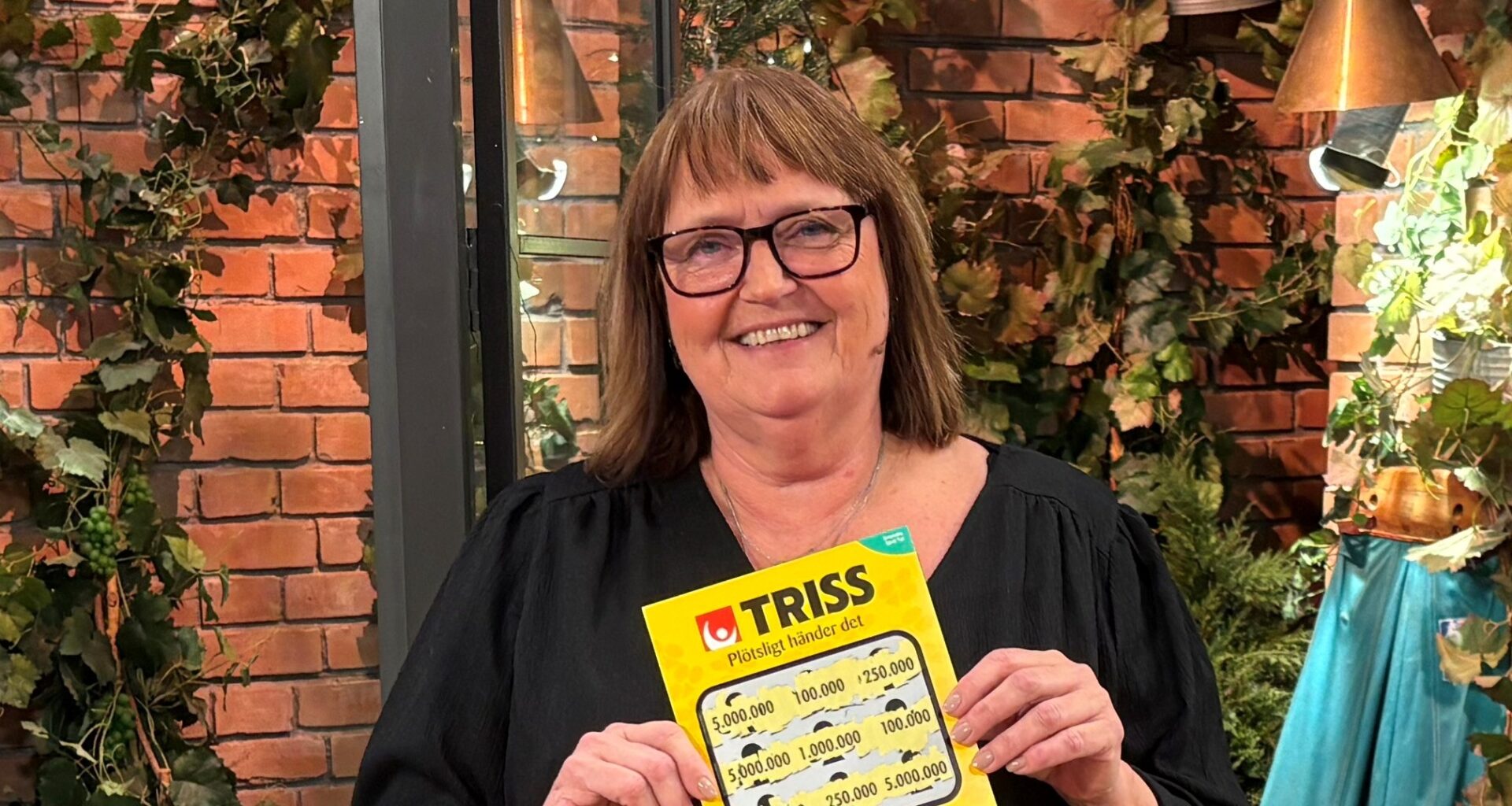 Person håller en Trisslott framför en dekorativ vägg med växter i bakgrunden.