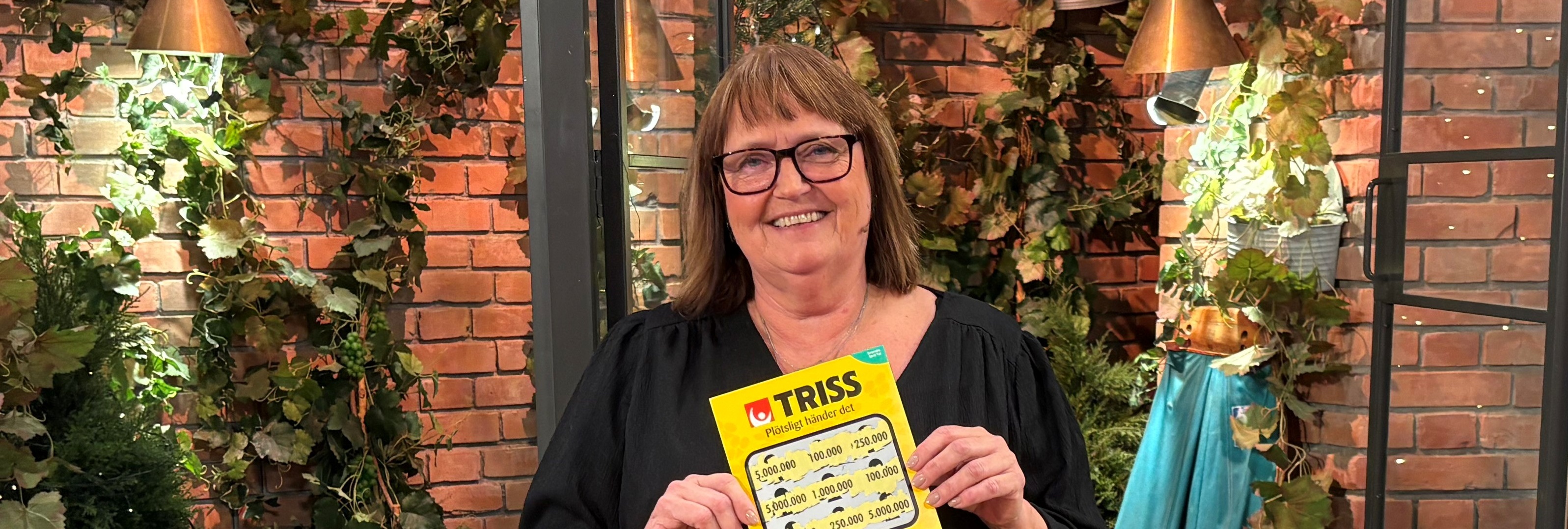 Person med Trisslott Person håller en Trisslott framför en dekorativ vägg med växter i bakgrunden.