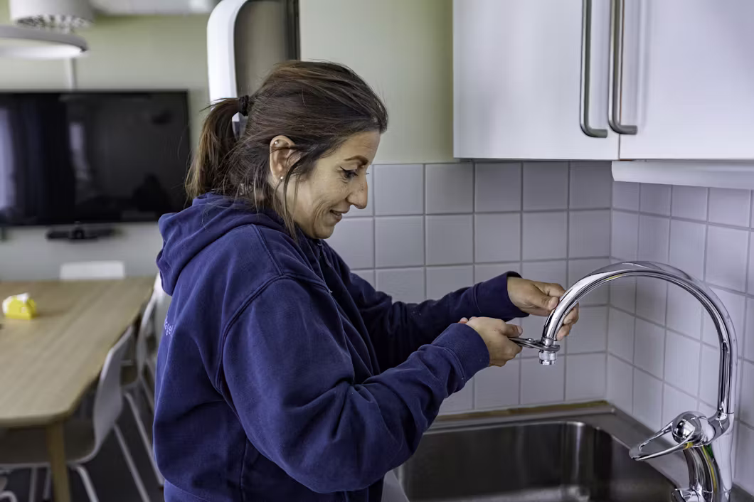 En kvinna i blå huvtröja justerar kranen i ett kök, står vid en diskbänk i rostfritt stål och kaklat stänkskydd.