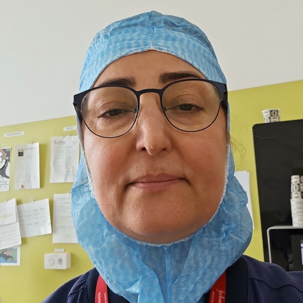 En person med glasögon, en blå engångsmössa och ett blått skäggskydd står inomhus framför en gul vägg med papperslappar och staplade muggar i bakgrunden.