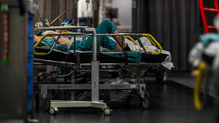 En patient ligger på en sjukhussäng i en vårdmiljö, omgiven av medicinsk utrustning.
