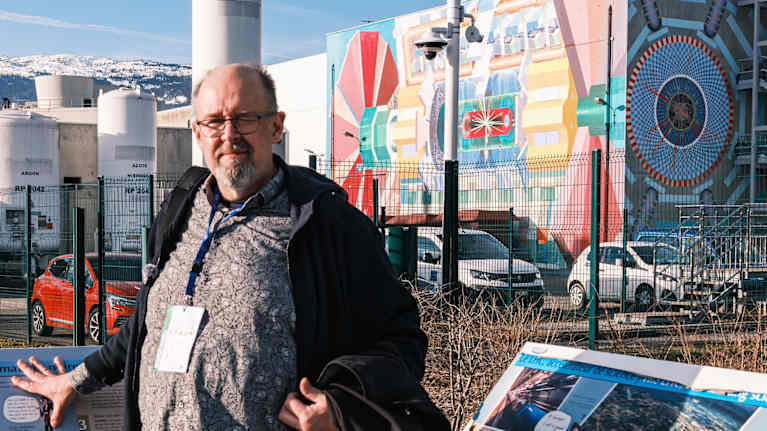 Marcus Rosenlund, en skäggig man med glasögon står framför en industriell anläggning med färgglada muralmålningar ,