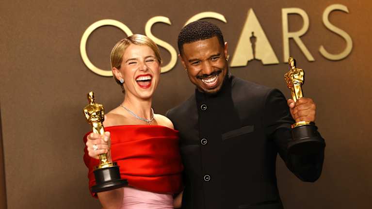 Finklädda Jessie Buckley och Michael B. Jordan ler stort medan de håller i guldiga Oscarsstatyetter. I bakgrunden syns Oscars logo på en svart vägg.