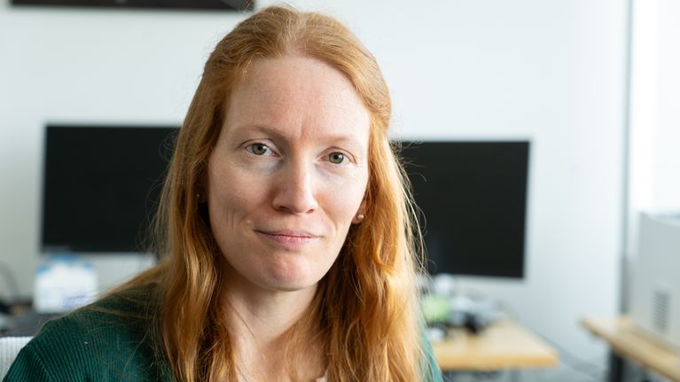 Personporträtt på kvinnlig läkare med långt rött hår och grön kofta, i bakgrunden hennes mottagningsrum.