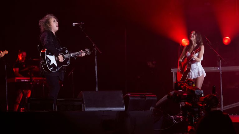 Två musiker står på en scen med gitarrer under en konsert med röda scenlampor i bakgrunden.