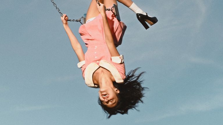 Olivia Rodrigo iklädd i rosa klänning och svarta skor gungar upp och ner mot en blå himmel.
