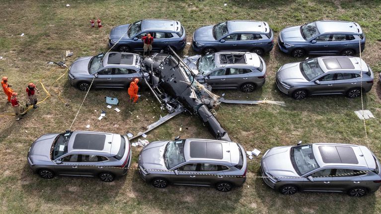 En kraschad helikopter ligger på marken omgiven av flera bilar; personer i orange kläder står intill vraket.
