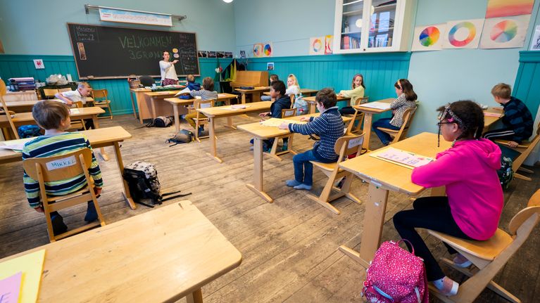 Elever sitter vid gamla träbord i ett klassrum med en lärare vid tavlan.