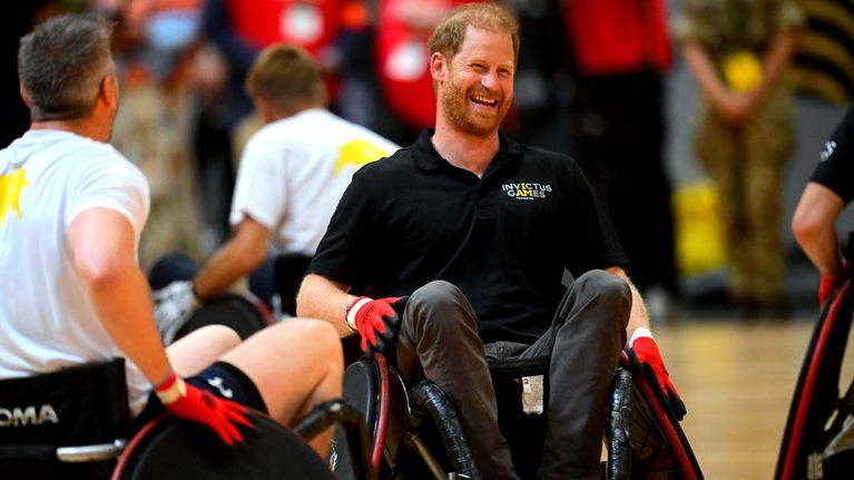 Prins Harry sitter i en en sportrullstol i en idrottshall och ler stort, omgiven av andra idrottare i rullstol.