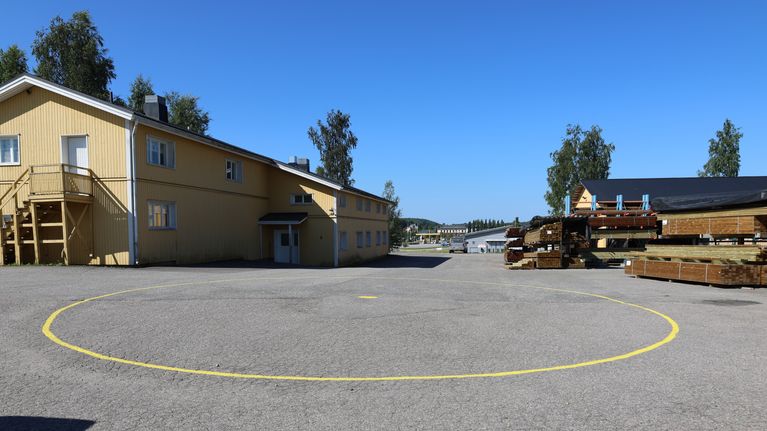 En asfalterad gårdsplan med en gul cirkel på marken, intill en gul träbyggnad och staplade träreglar.