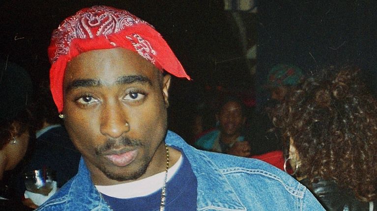 Tupac Shakur i en blå tröja, jeansjacka och röd bandana tittar in i kameran. Han visar mittfigrarna mot kameran.