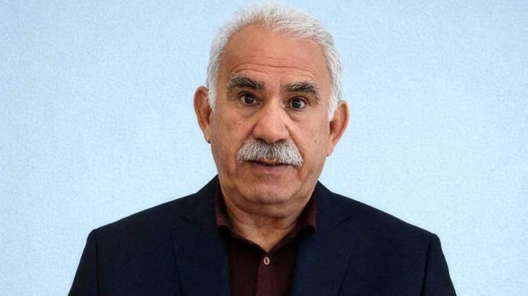 Abdullah Öcalan sitter i kavaj och mörk skjorta vid ett bord med en bunt papper framför sig, ett papper i handen och ser in i kameran.
