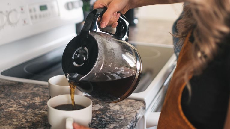 En kvinna häller upp bryggkaffe från en kaffekanna i en vit kopp som står på en köksbänk.