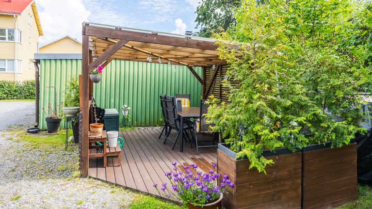 En uteplats med trädäck, matbord och stolar står under ett träpergola bredvid grön vägg och blomlådor.