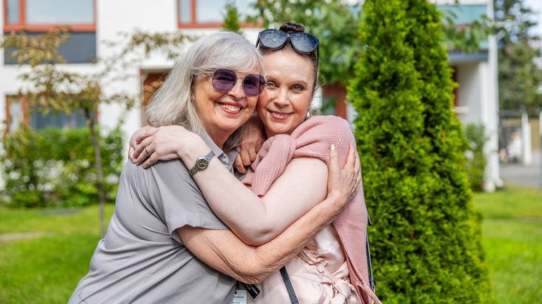Två kvinnor står ute i en trädgård och kramar varandra framför ett vitt bostadshus.