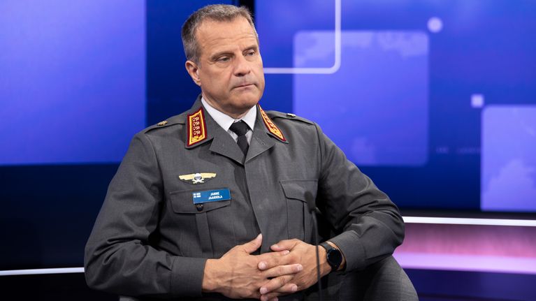 En man i militäruniform sitter i en tv-studio med blå bakgrund och digitala rektanglar.