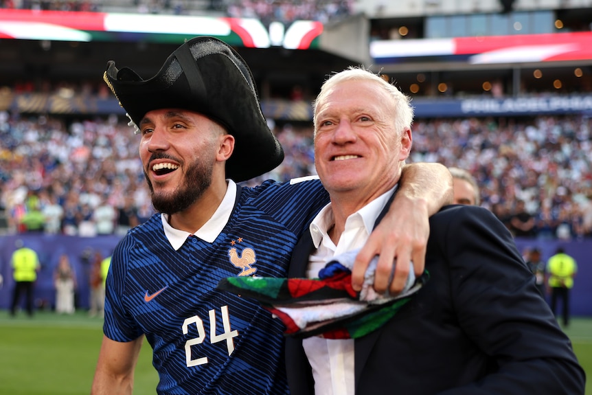 Rayan, wearing a black hat, has his arm around Didier. Both smile.