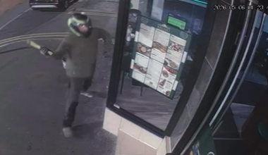 Photo of a man smashing up the windows of a turkish restaurant. he is wearing a bike helmet and a dark green coat.