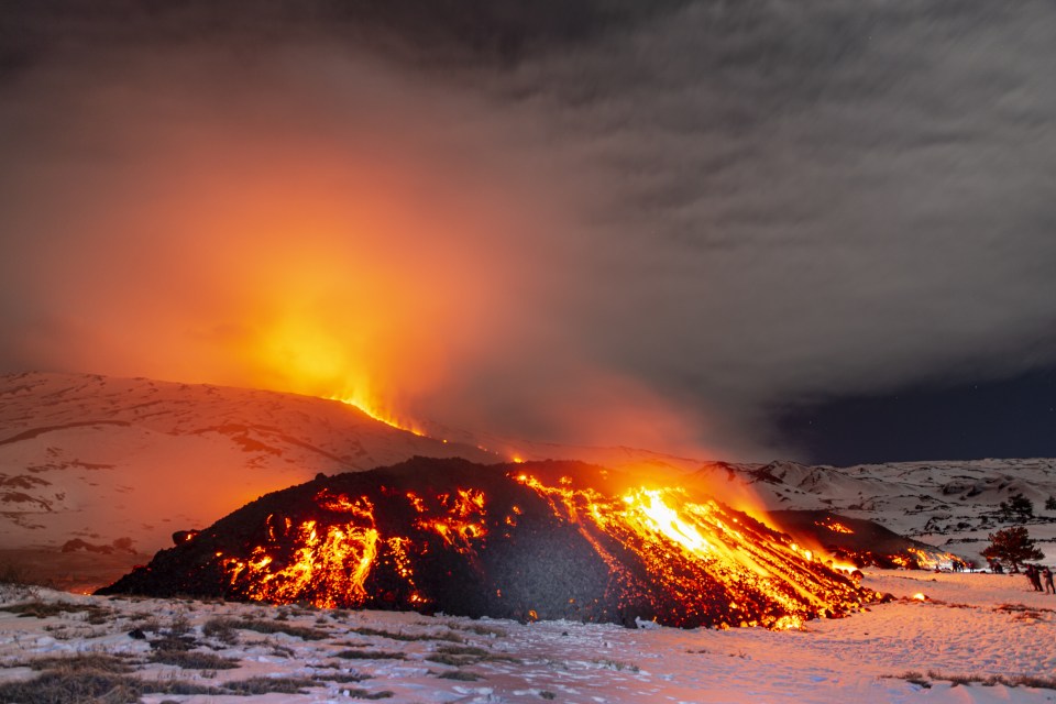 Lava flowing down Mount Etna at night, with tourists watching from a distance.