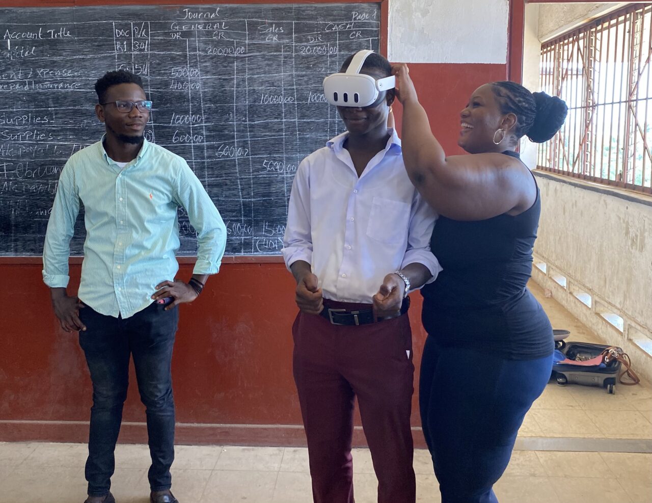 In a room with a chalkboard and bright-red walls, a woman stands next to a person wearing a white-colored device that covers the eyes and is strapped onto the head. A third person in a pastel green shirt looks on.