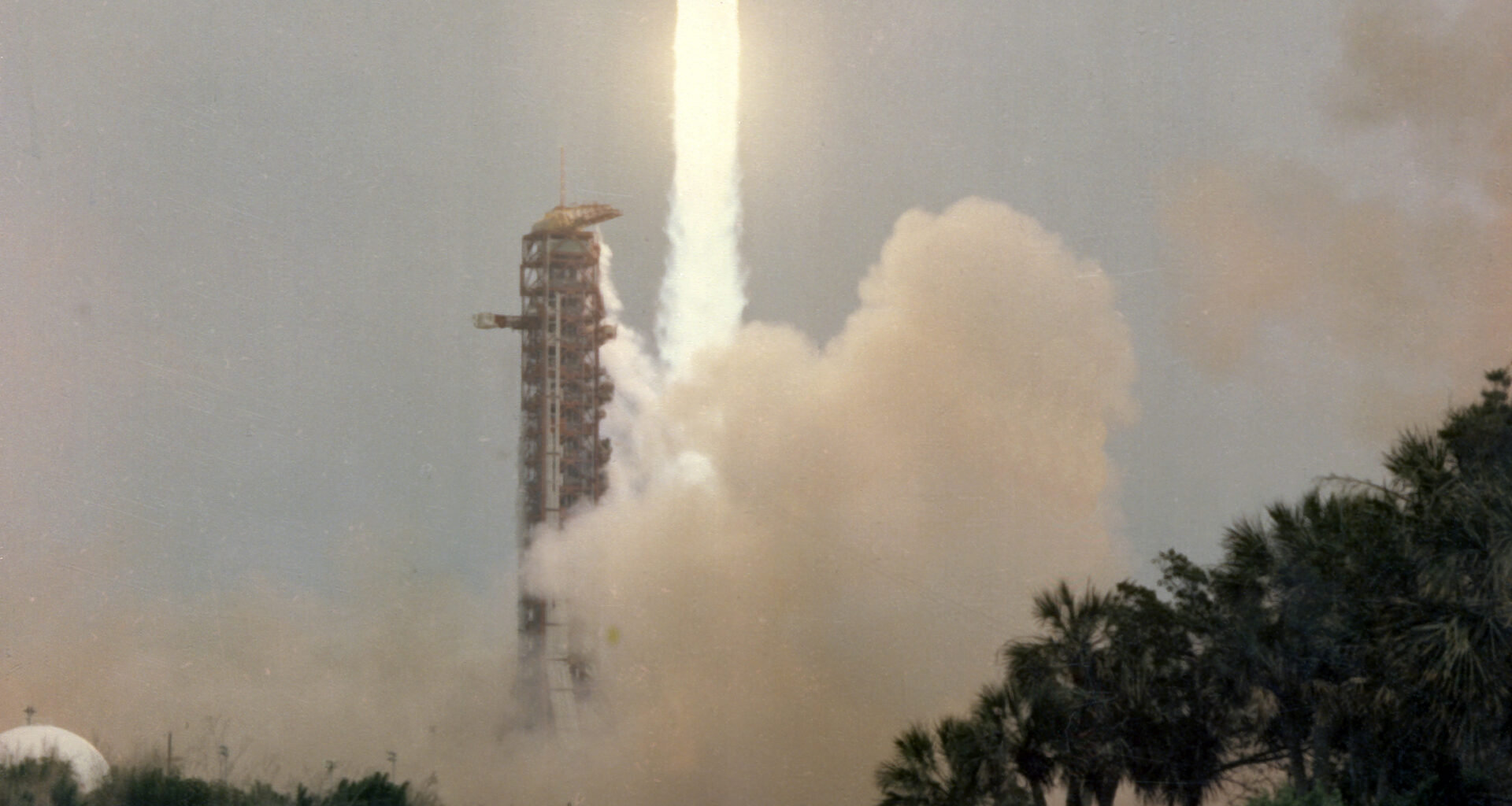 Apollo 13 launches upward, leaving a column of flames and light brown vapor behind it. Trees and vegetation frame the water at the bottom of the photo. Outlines of small animals can be see in the water. At bottom left is written "Apollo 13, launch" in capital letters. At bottom right is a series of letters and numbers labeling the photo.