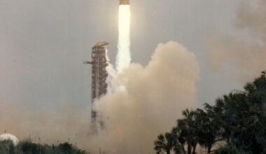 Apollo 13 launches upward, leaving a column of flames and light brown vapor behind it. Trees and vegetation frame the water at the bottom of the photo. Outlines of small animals can be see in the water. At bottom left is written "Apollo 13, launch" in capital letters. At bottom right is a series of letters and numbers labeling the photo.