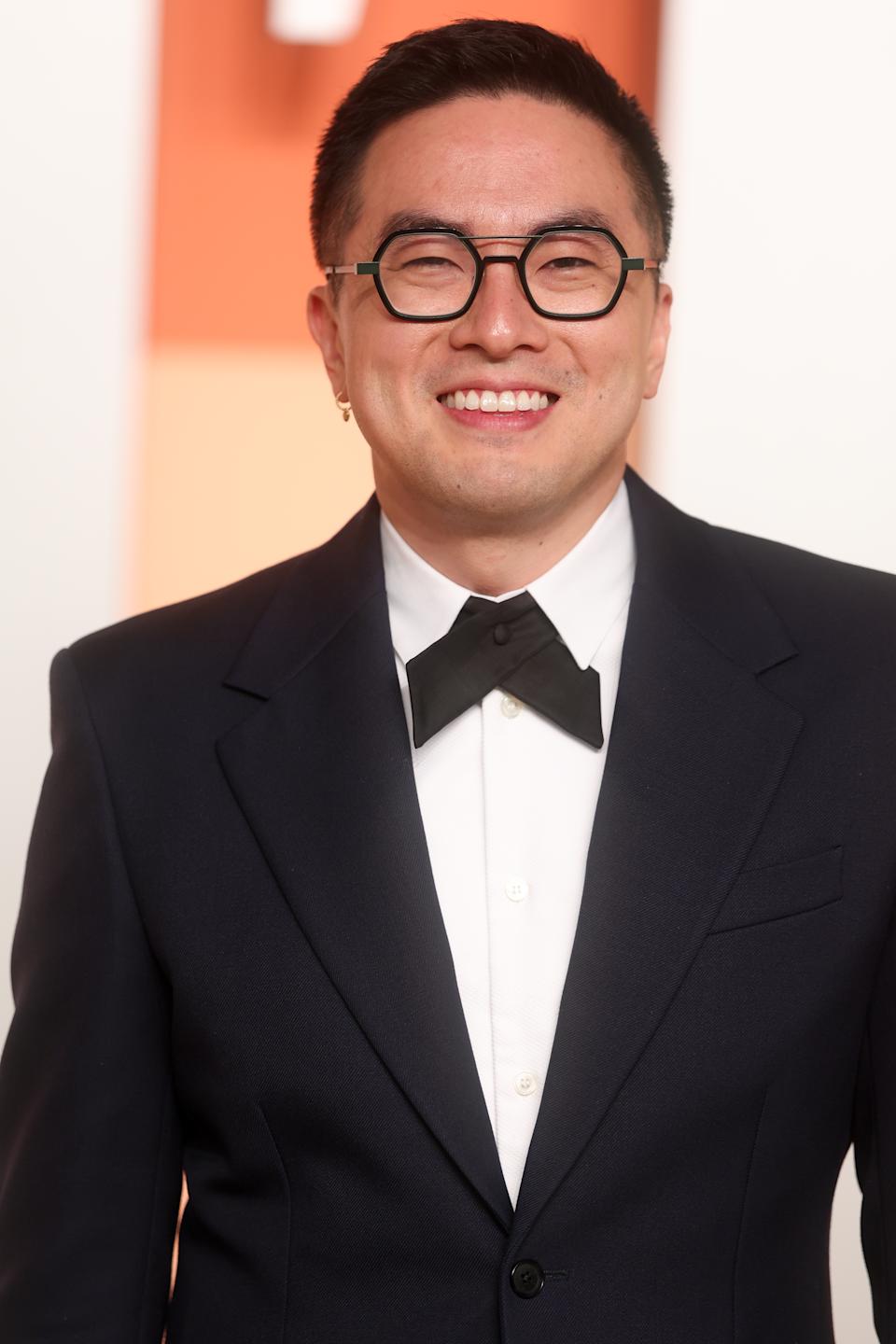 Bowen Yang in a formal suit with a bow tie stands smiling on a red carpet