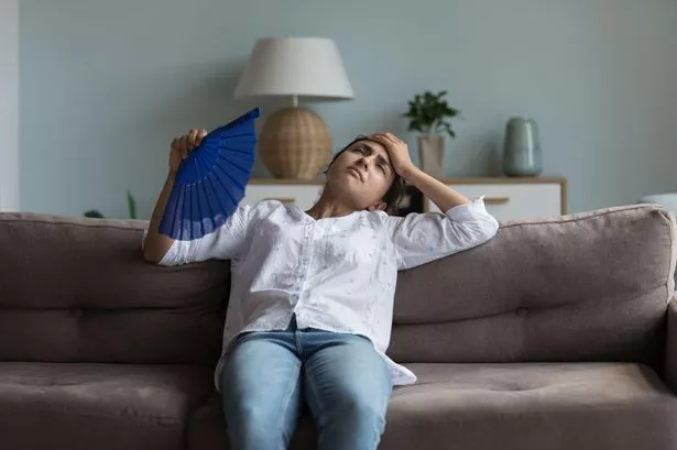 This is a photo of a young woman struggling in the heat sat on a sofa fanning herself. 