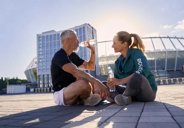 This is a photo of a middle-aged couple sat on the ground after a run drinking water.