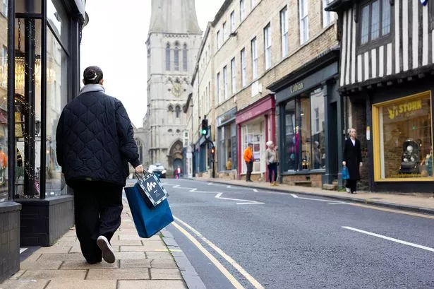 Woman with two shopping bags walking across pavement with shops pictured on the opposite side of the road 