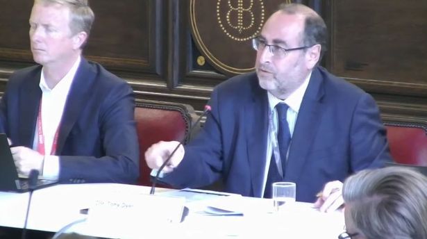 Man in blue suit wearing glasses sat at table speaking into microphone at council meeting