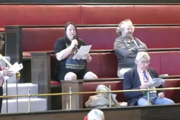 Woman in black T-shirt and shorts sat on red benches in public gallery at council chamber speaking into microphone
