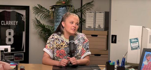 JoJo Siwa, in a colourful top with cartoons on, sat in a podcast studio with a microphone in front of her. 
