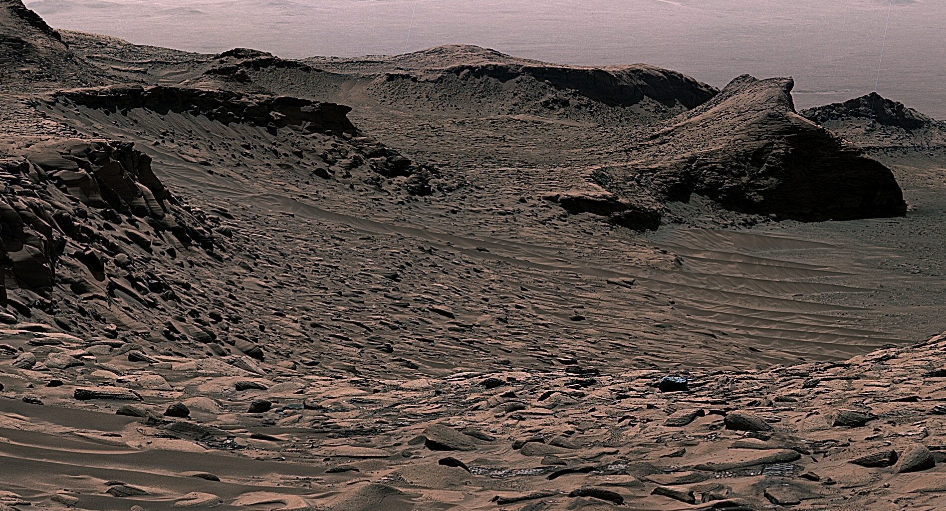 An image of Mars shows rocks and small sand dunes, with a larger mountain range in the background. Two wheel tracks in the foreground show Curiosity rover's path before it captured the image.